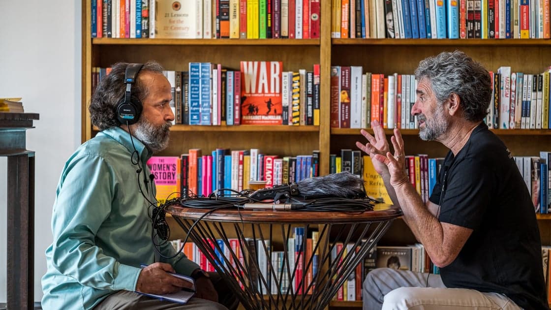 Two men in an interview setup in front of a bookshelf, one wearing headphones and taking notes, the other gesturing while speaking